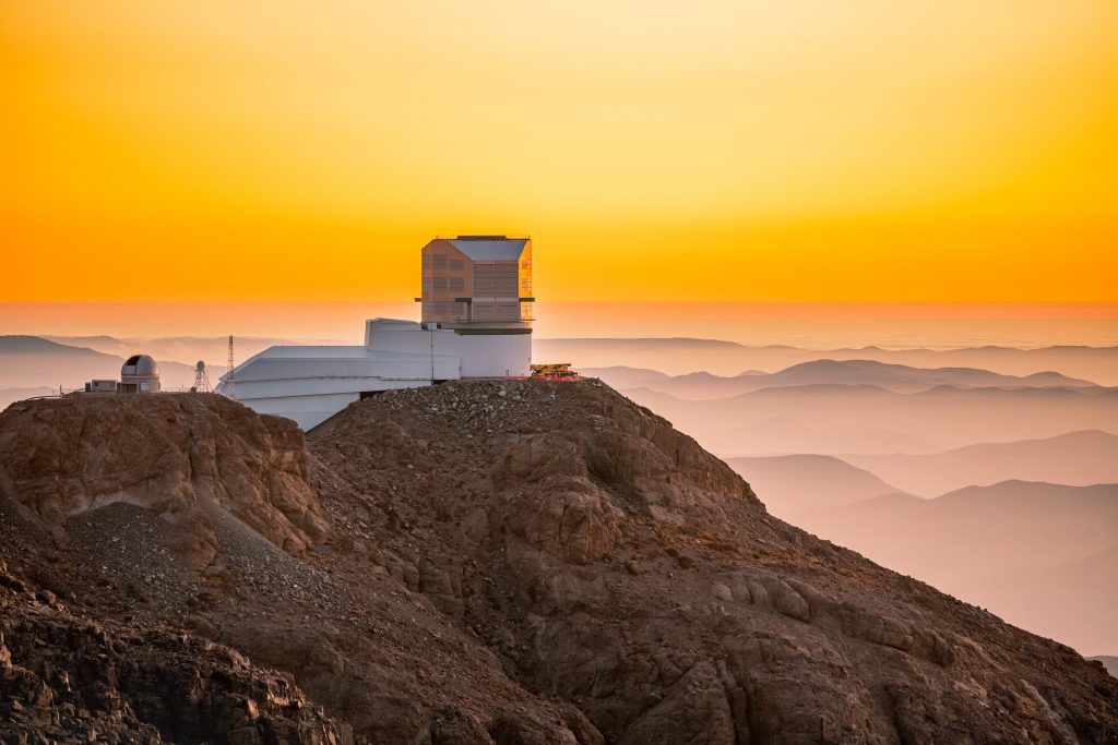 The Rubin Observatory seen on top of a mountain above cloud level at sunset. The sky is bright orange and clouds can be seen in the distance lower than the observatory.